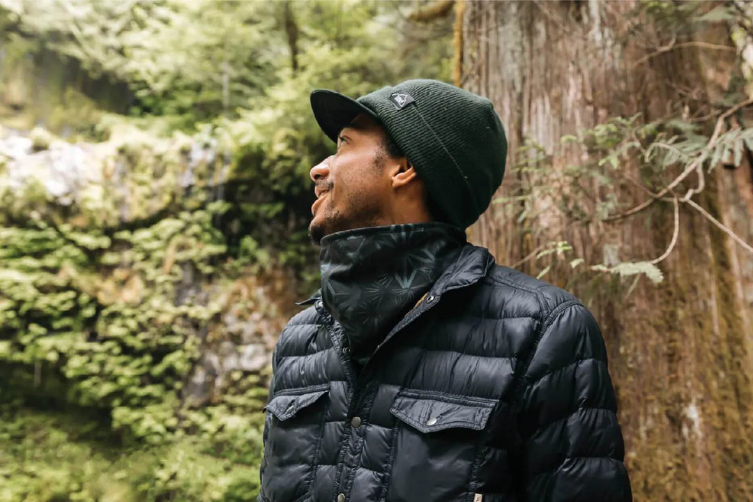 A man wearing a black jacket and topone neckgaiter in the woods.